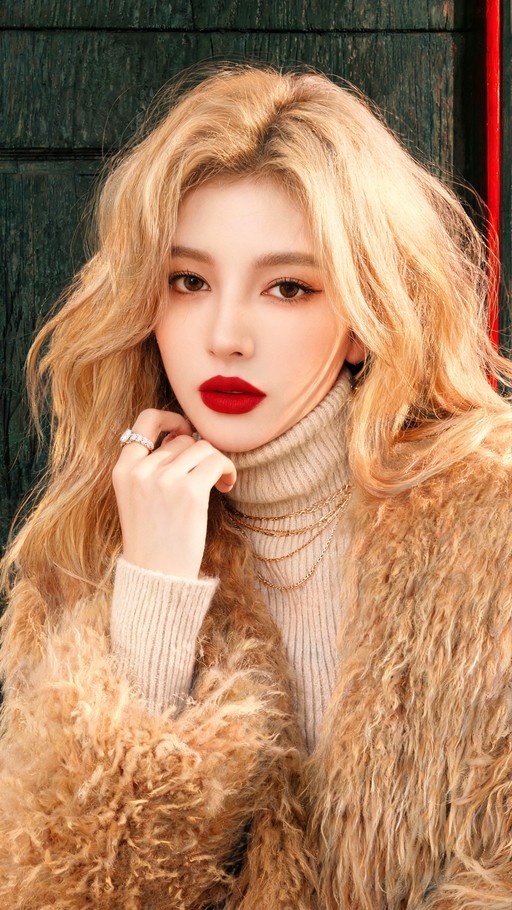 A blonde woman with wavy hair wearing a beige turtleneck and brown fur coat, looking at the camera with red lipstick.