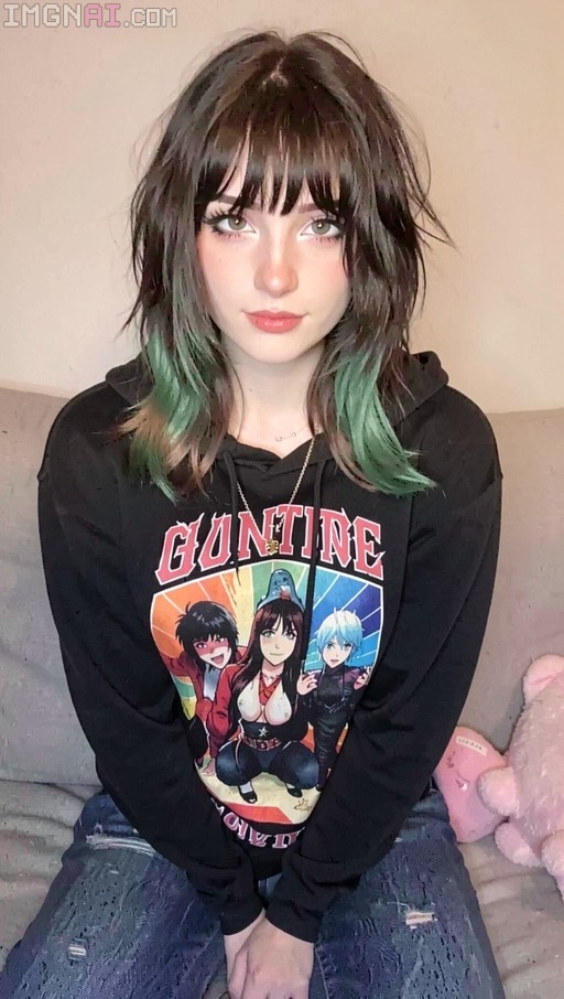 A young woman with dark hair and green tips sits on a couch wearing a black Guntire anime hoodie and ripped blue jeans.