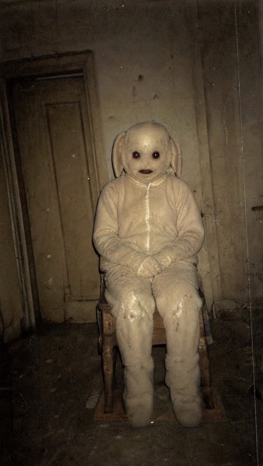 A full-body shot of a pale figure in a textured white bunny costume sitting on a wooden chair in a dim room, looking directly at the viewer with dark eyes.