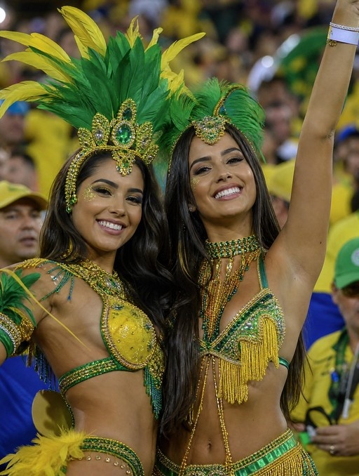 Two smiling women in elaborate green and gold feathered carnival costumes pose together with one raising her arm in celebration against a blurred crowd background.