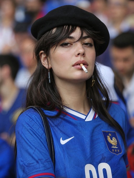 A young woman with dark hair wearing a black beret and a blue France soccer jersey smokes a cigarette in a crowded stadium.