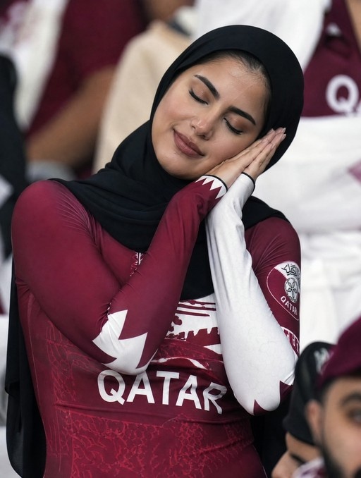 A realistic photograph of a young woman wearing a black hijab and a maroon Qatar sports jersey, sleeping with her eyes closed and head resting on her hands in a crowded stadium.