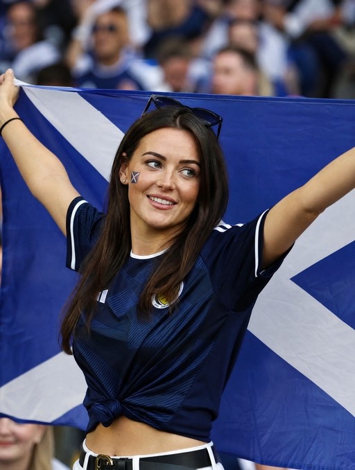 A young woman with long dark hair smiles while holding a large Scottish Saltire flag behind her head at a soccer match, wearing a tied blue jersey and sunglasses perched on her head.
