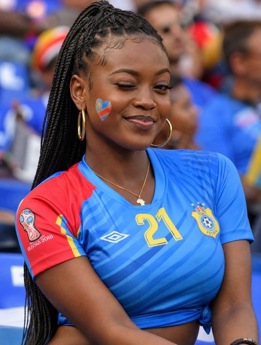 A smiling DR Congo fan at the 2018 World Cup wearing a blue jersey with red sleeves, gold hoops, and a heart-shaped flag sticker on her cheek.