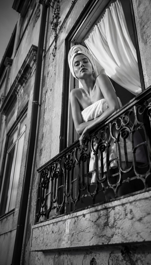 Black and white photograph of a woman wrapped in towels leaning against an ornate iron balcony railing in front of a window with curtains.
