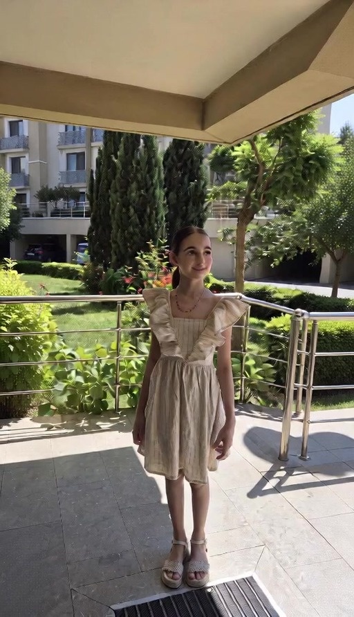 A smiling woman with dark skin and a black ponytail stands outdoors in a yellow dress holding a suitcase near trees and buildings.