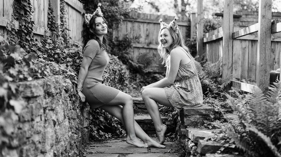 Two smiling women wearing cat ears sit barefoot on stone steps in a garden.