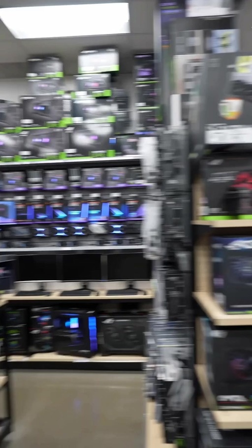A realistic video shows a man with grey hair and glasses holding a microphone in a computer hardware store, interviewing shoppers about rising graphics card prices.