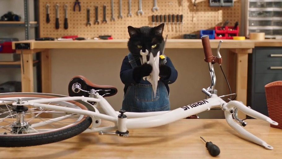 A black cat wearing denim overalls stands at a workbench holding a wrench and screwdriver while looking at bicycle instructions with a confused expression before throwing the tools down in frustration.