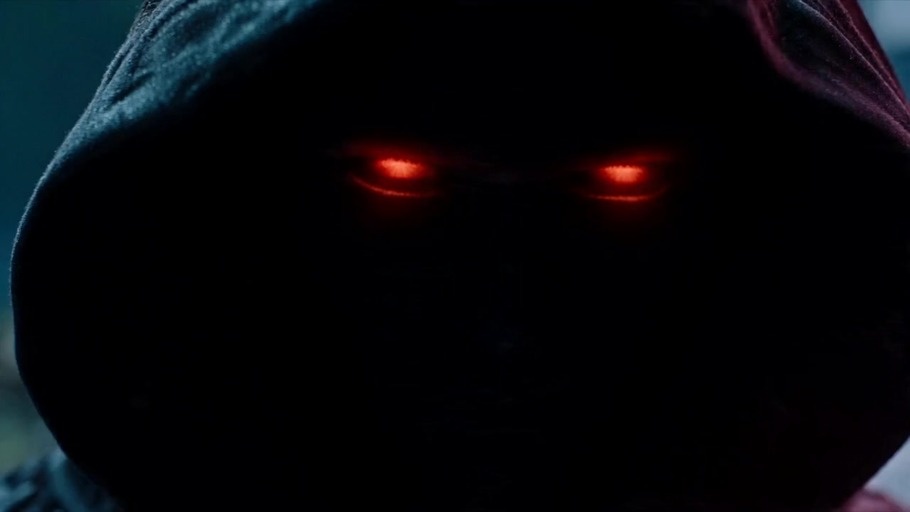 A dark hooded character with glowing red eyes stands opposite a figure in a red hoodie with X-shaped eyes before the red figure fires a bright energy beam that explodes a wooden building.