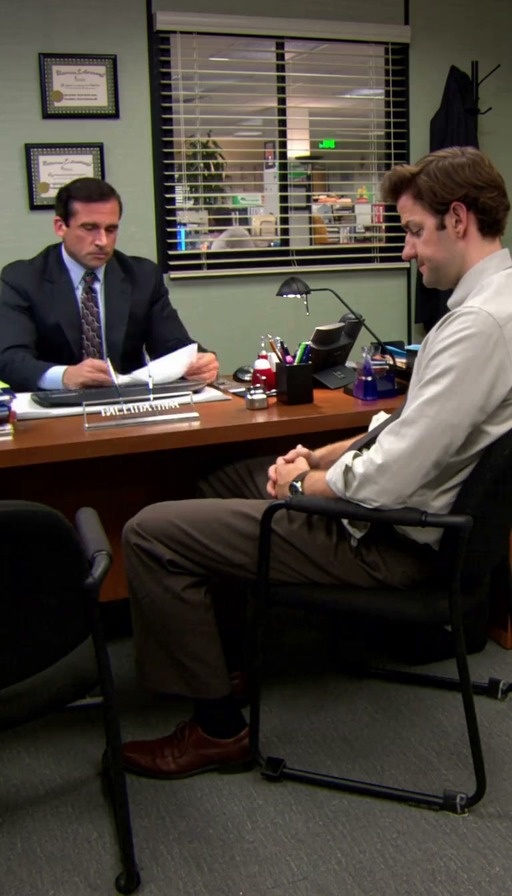 Michael Scott sits at his desk in a suit holding papers while Jim Halpert sits opposite him looking guilty during a serious conversation.