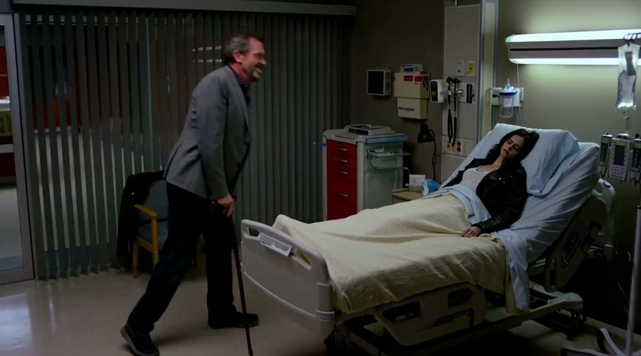 Dr. House leans over Jessica Jones in a hospital bed before she punches him.