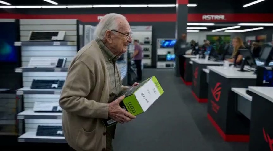 An elderly man with glasses and a beige cardigan holds a large graphics card box while standing at an electronics store checkout counter.