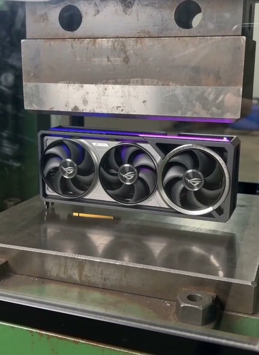 A triple-fan ASUS ROG graphics card is slowly crushed by a large industrial hydraulic press, with its fans and casing deforming under the pressure.