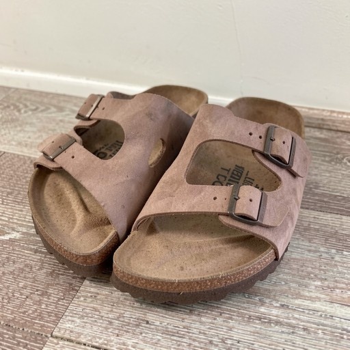 A pair of brown two-strap leather sandals with cork soles resting on a wooden floor.