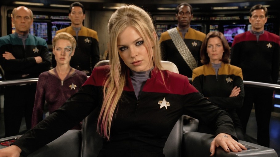 Avril Lavigne sitting in a command chair on a spaceship bridge wearing a black and red uniform with pink highlights in her blonde hair.