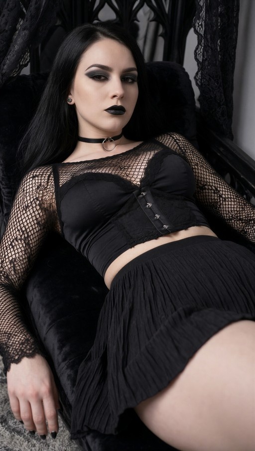 A woman with long black hair and dark lipstick wearing a black fishnet top and pleated skirt reclines on a chair while looking at the viewer.