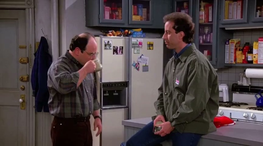 Jerry Seinfeld sits on the kitchen counter while George Costanza, wearing glasses and a plaid shirt, stands nearby holding a mug and gesturing during their conversation.