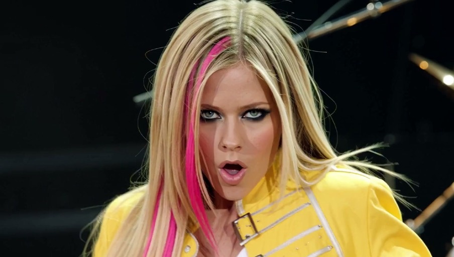 Avril Lavigne with blonde hair and pink streaks wearing a yellow jacket and white pants performing on stage with a microphone stand.