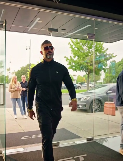 A muscular man in sunglasses and black athletic wear walks into a computer store and points at a high-end gaming PC on the counter.