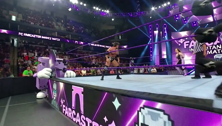 A blonde female wrestler in a black and purple outfit stands over a fallen opponent before being grabbed from behind by a large grey-skinned wrestler who applies a chokehold.