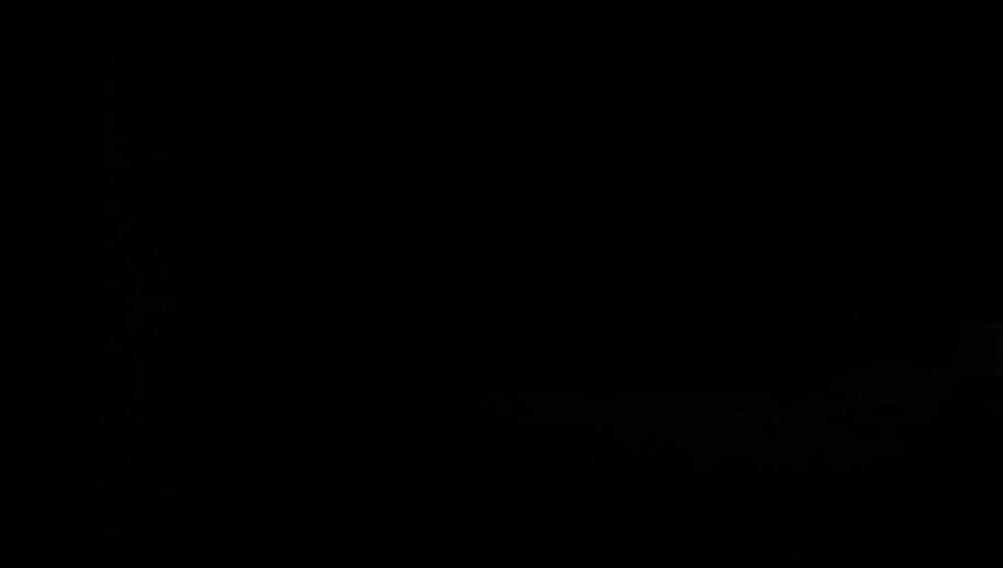 A purple-haired anime boy in a black shirt walks into a dark room and falls onto the bed while a small cat wearing a witch hat jumps up next to him.