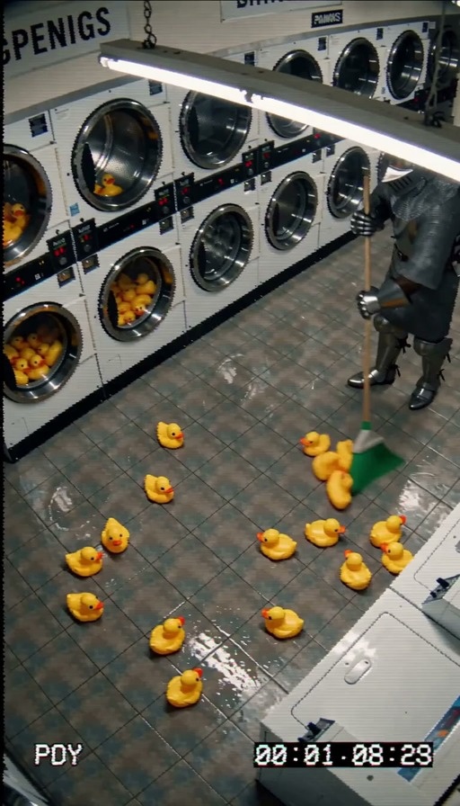 A person in full medieval armor sweeps yellow rubber ducks into a pile on the tiled floor of a laundromat, viewed through a grainy security camera lens.