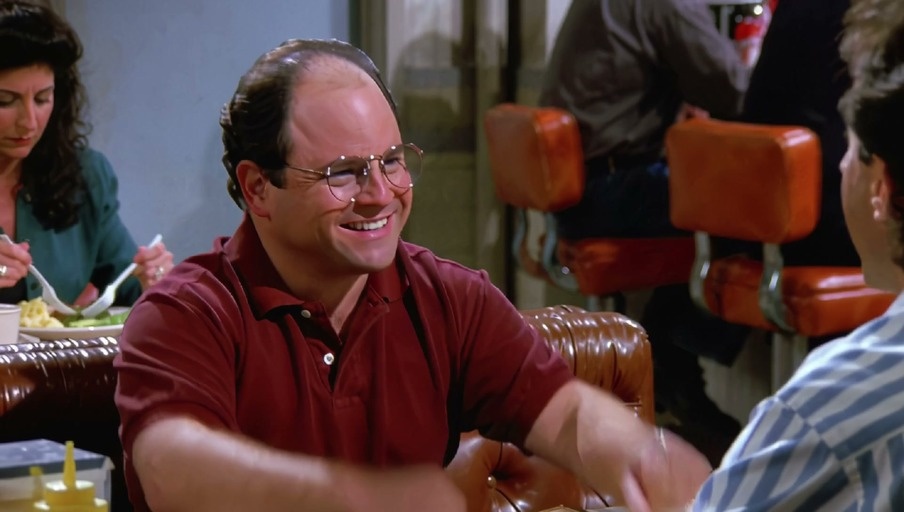 George Costanza in a red shirt and glasses gestures excitedly while talking to Jerry Seinfeld at a diner table.