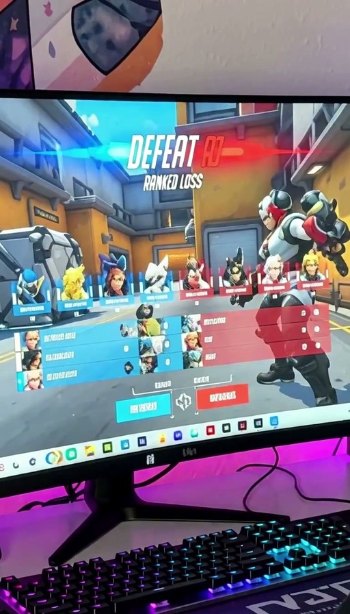 A pink-haired woman with cat ears sits at her computer playing Overwatch 2 and reacts to a ranked loss.
