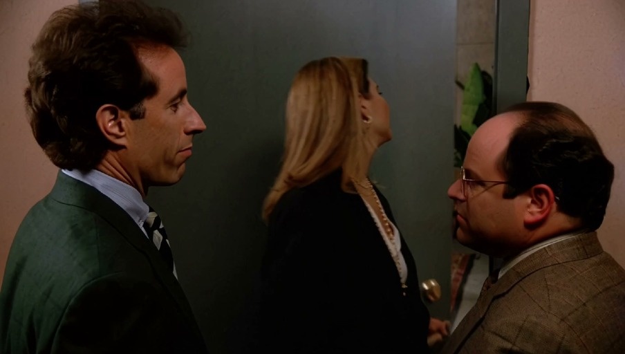 Jerry Seinfeld and George Costanza stand in a doorway watching executives, including one in an alien mask, stand up to greet them.