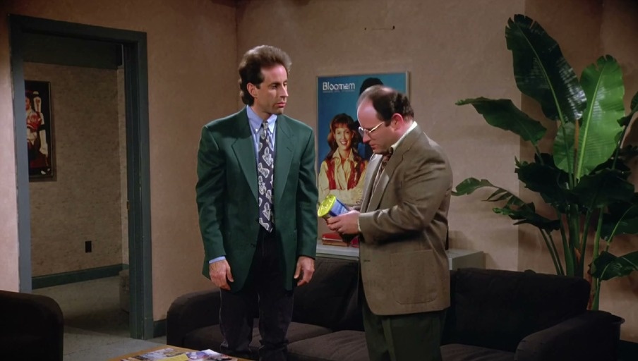 George Costanza wearing a green jacket holds up a can of Spam while standing next to Jerry Seinfeld in a living room.